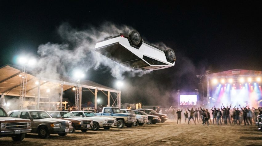 Carro branco angular dando cambalhota no ar sobre uma fileira de veículos, em um evento noturno no Texas (ilustração criada com IA)